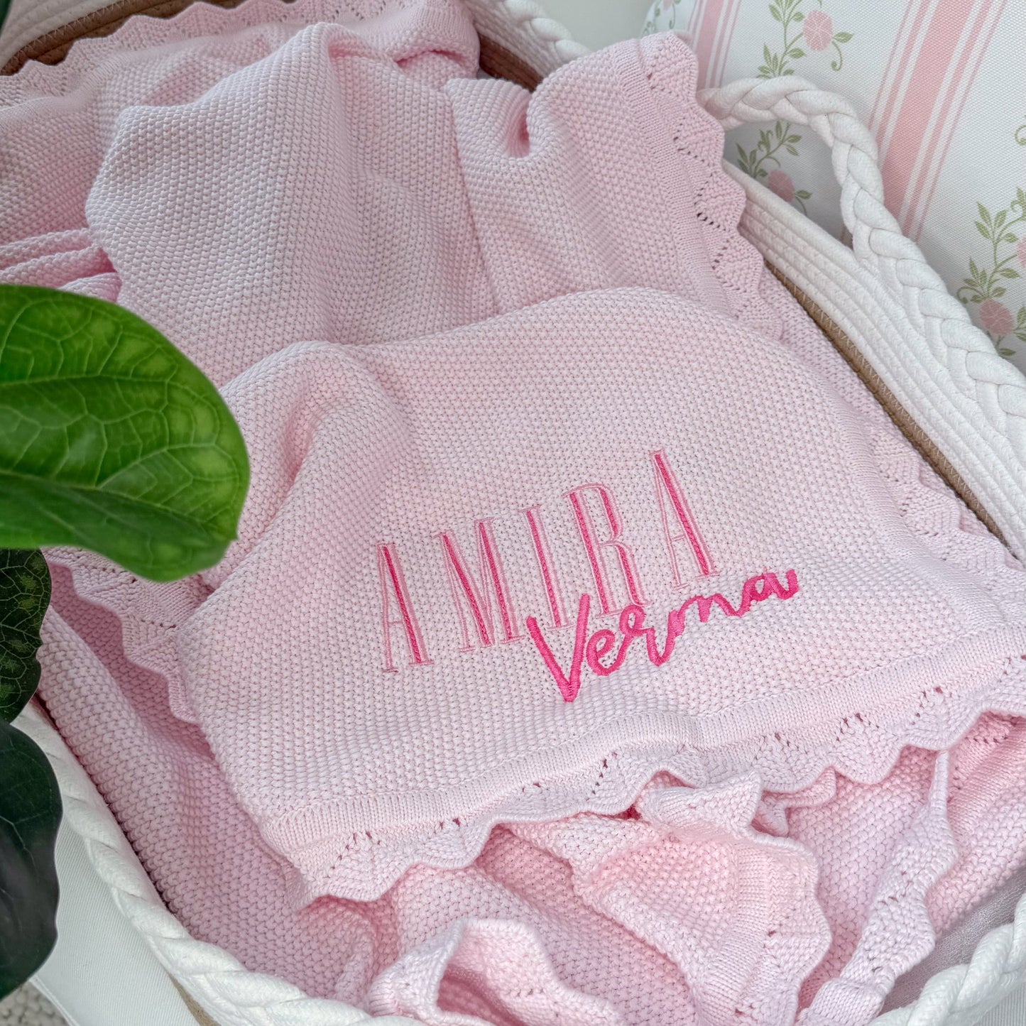 A pink textured baby blanket with ruffled edges and personalized embroidery reading 'Amira Verma' in pink, displayed inside a white woven basket next to a green plant leaf.