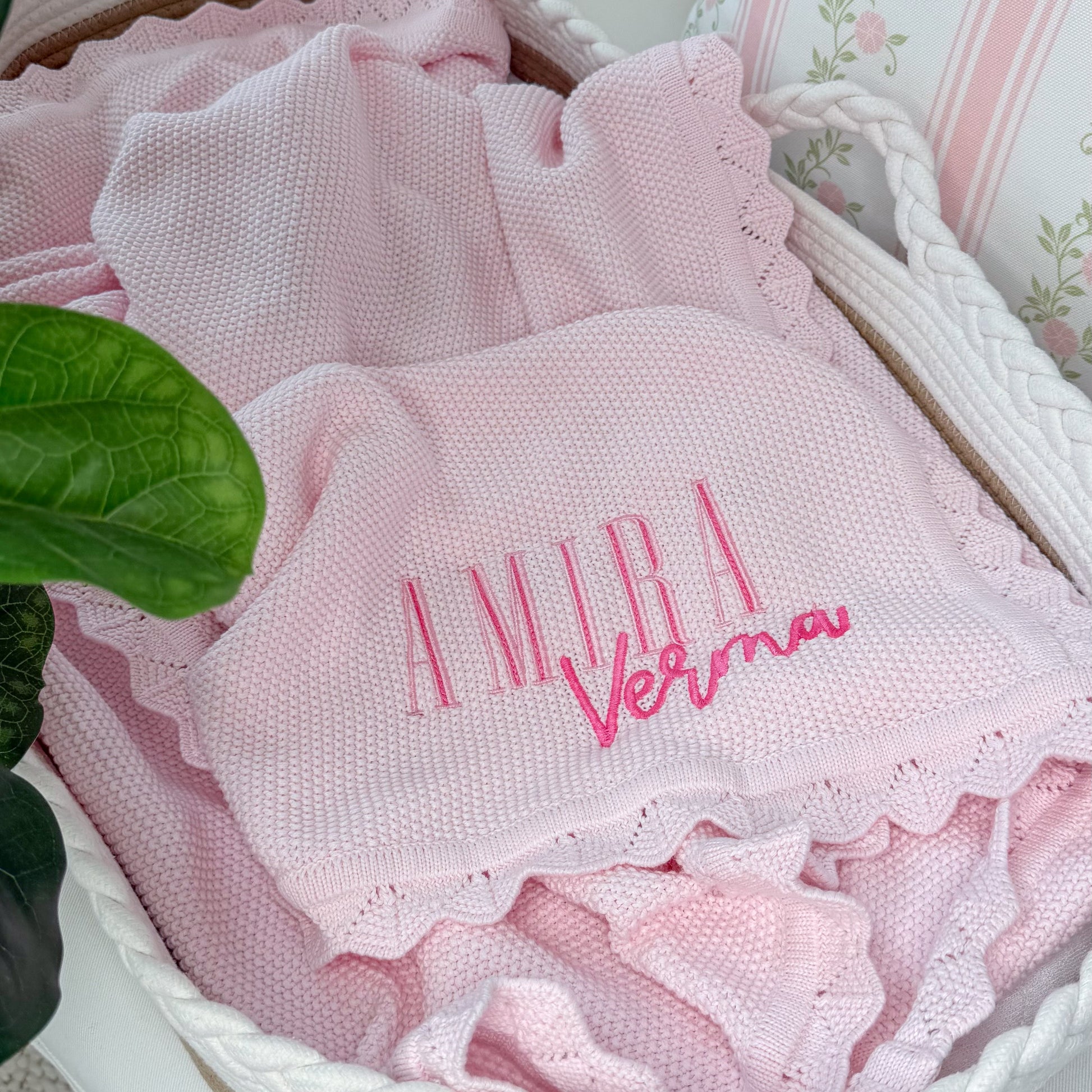 A pink textured baby blanket with ruffled edges and personalized embroidery reading 'Amira Verma' in pink, displayed inside a white woven basket next to a green plant leaf.